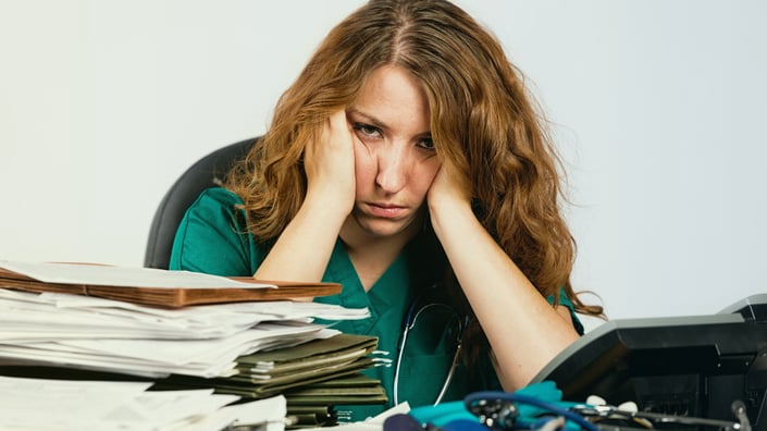 Stressed healthcare credentialing manager overwhelmed with paperwork, sitting at a cluttered desk with a computer displaying multiple credentialing applications.